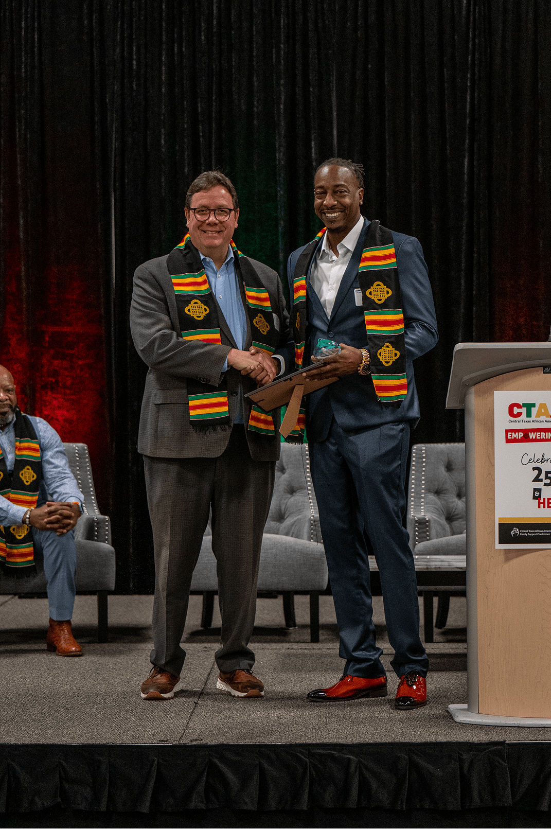 Two men in African-inspired stoles shake hands during a formal event, celebrating achievements in a supportive atmosphere. A third man sits in the background.