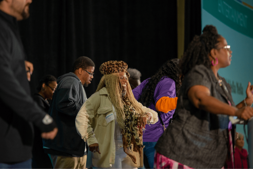 A group of people joyfully dances on stage at an event, showcasing community engagement and celebration. The focus is on a woman in a leopard-print hat, smiling as she participates.