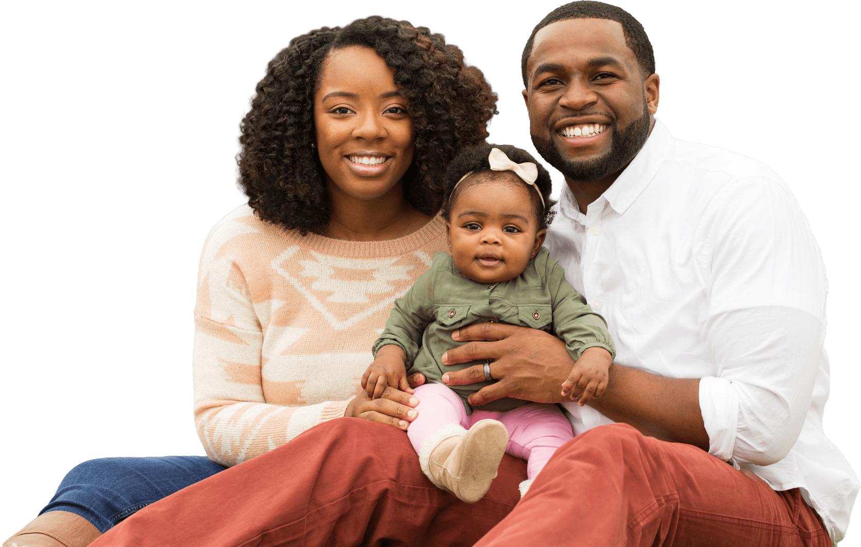 A smiling couple sits together with their baby girl, showcasing a warm family moment. They wear casual, coordinated outfits against a neutral background.